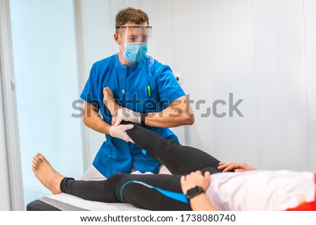 A physiotherapist with mask and screen giving a massage to an ankle. Physiotherapy with protective measures for the Coronavirus pandemic, COVID-19. Osteopathy, therapeutic chiromassage