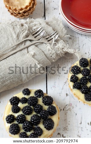 two homemade blackberries tart with pastry cream on wooden table with cloth, fork and little plate with tart waffles
