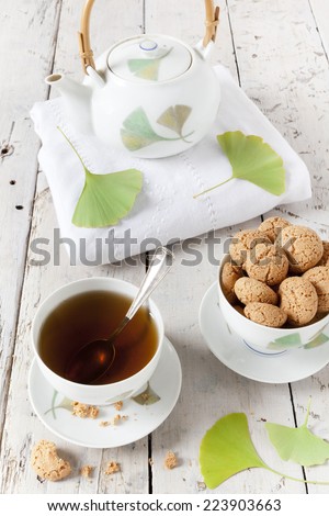 tea on cup with teapot and amaretti sweets on white table with gingo biloba leaves