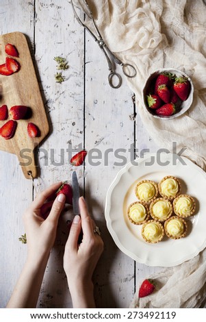 view from above of hands preparing little tartlets with pastry cream and sliced strawberries on rustic table