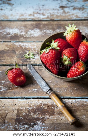 small bowl of fresh whole strawberries on a rustic grungy wooden table with knife and one bitten strawberry
