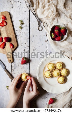 view from above of hands preparing little tartlets with pastry cream and sliced strawberries on rustic table