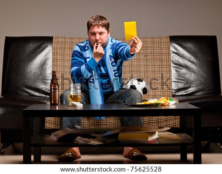 soccer fan is sitting on sofa with beer and showing yellow card at home