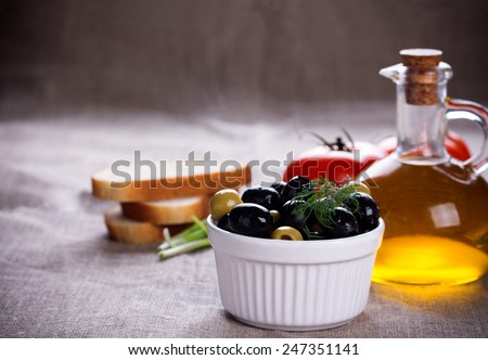 Bread, red tomatoes, black and green olives in white bowl and small bottle of olive oil on jute fabric.
