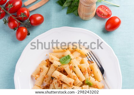 Plate of penne pasta with bread crumbs, basil and cherry tomatoes on blue tablecloth.