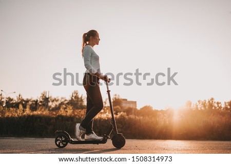 Young beautiful girl riding an electric scooter in the summer on the street, against the sunset, there is free space for advertising against the sky