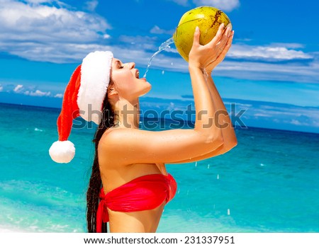 Young beautiful woman with long black hair in red bikini, dressed in red Santa Claus hat is drinking coconut water on tropical beach. Christmas and New Year on the tropical coast.