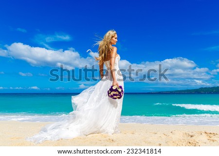 beautiful blond fiancee in white wedding dress with big long white train and with wedding bouquet stand on shore sea