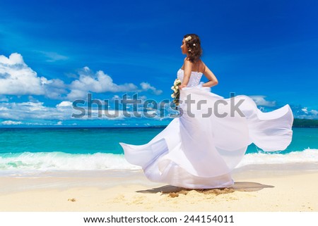 Beautiful brunette bride in white wedding dress with big long white train and with wedding bouquet stand on shore sea. Tropical sea in the background. Summer vacation concept.