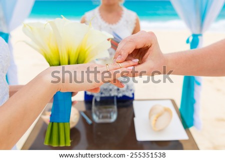 Groom giving an engagement ring to his bride under the arch decorated with flowers on the sandy beach. Wedding ceremony on a tropical beach in blue. Wedding and honeymoon concept.