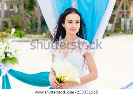 Wedding ceremony on a tropical beach in blue. Happy bride under the wedding arch decorated with flowers on tropical sand beach. Wedding and honeymoon concept.
