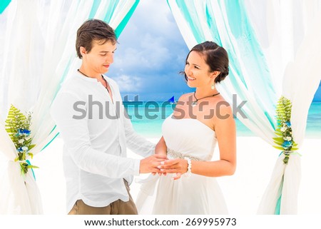 Bride giving an engagement ring to her groom under the arch decorated with flowers on the sandy beach. Wedding ceremony on a tropical beach in blue. Wedding and honeymoon concept.