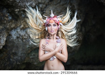 Fantasy photo of a young beautiful girl with curly hair and ornaments made of shells standing on a rock tropical island. Summer vacation concept.
