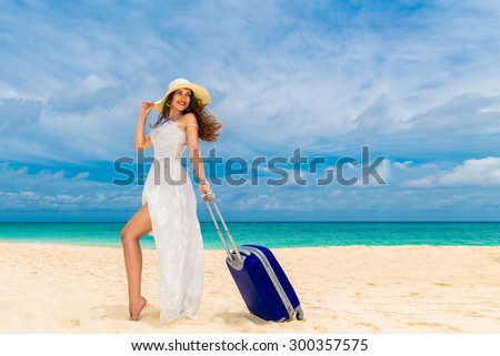 Beautiful young woman in white dress and straw hat with a suitcase on a tropical beach. Blue sea in the background. Travel concept.