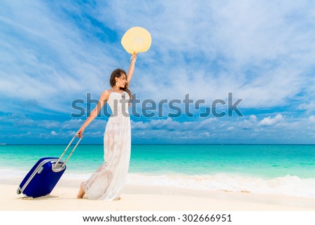 Beautiful young woman in white dress and straw hat with a suitcase on a tropical beach. Blue sea in the background. Travel concept.