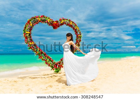 Wedding ceremony on a tropical beach. Happy bride under the wedding arch in the form of heart decorated with flowers on tropical sand beach. Wedding and honeymoon concept.