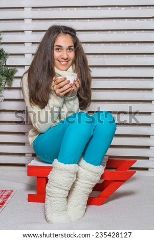 Beautiful and happy girl with gifts near a Christmas tree wishes everyone a Merry Christmas and Happy New year sits on a sled and drinking cappuccino