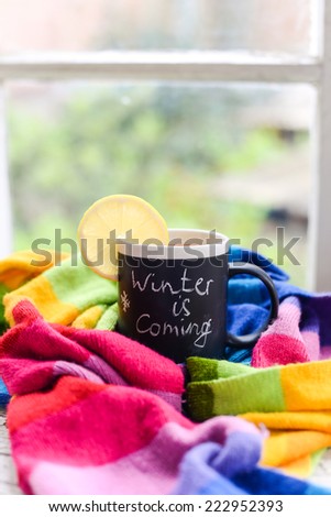 A mug with chalk writing Winter is coming with tea and lemon slice on it, wrapped in a multicolor rainbow scarf
