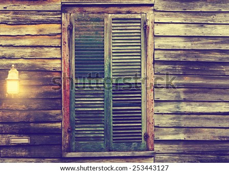 Old rustic vintage antique dilapidated run down leaning house home building structure with green closed shut shutters over window and porch light lantern glowing turned on