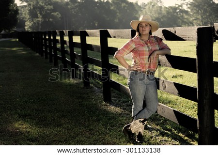Cowgirl lady woman female wearing cowboy hat and flannel shirt with jeans leaning on country rural fence by a horse pasture paddock looking confident happy serene smart alone waiting watching patient