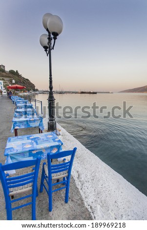 Some tables and chairs that belong to a tavern, in a row, on a jetty of Ikaria island, Greece, port,  during sunset.