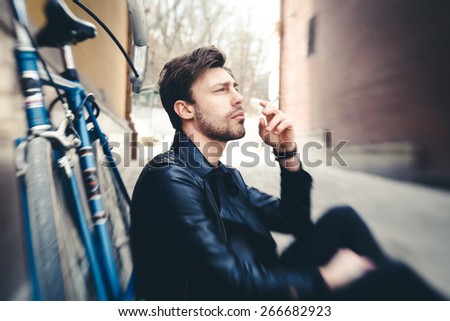 Close up fashion portrait of stylish handsome hipster guy sitting at city park near his fixed bike, lensbeybi, tilt-shift lens