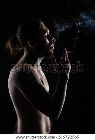 Portrait of a man with naked torso and tattooes. Dark and deep shadows. smokes in the studio