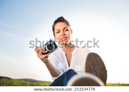 Beautiful girl sitting on the meadow with old vintage camera at sunset with soft blue sky. Copy space. Moment from the summer holiday. Bellow perspective.