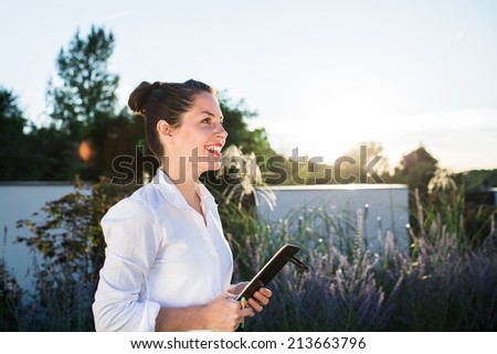 Young happy businesswoman or landscape architect in white shirt speaking with her client in the dark background with trees, bushes and the garden fence. Strong sunset backlit.