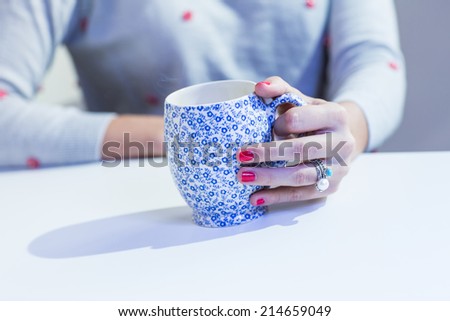 Young woman in grey sweater with red spots holding decorative cup of tea or coffee with smoke. Detail of the red painted nails. Color toned image. Cold filter.