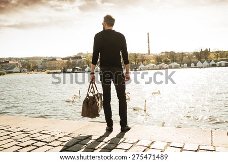Young stylish man with his bag is standing in front of river and looking in the city. Color toned image.