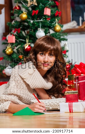 Beautiful young woman lying on carpet with a red pen writing a christmas card / Christmas presents in front of tree over living room