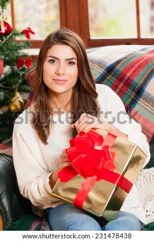 Happy young woman sitting on couch holding christmas present boxes next to christmas tree and fire place