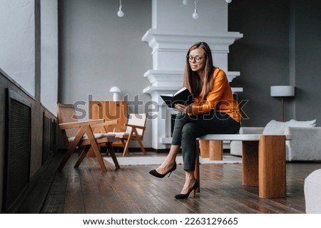 Cheerful young beautiful woman in orange blouse, black pants sitting with diary smiling looking at notebook, satisfied by business. Happy student girl reading book. Young housewife making store list Stok fotoğraf © 