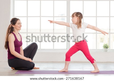 Young mother and daughter doing yoga exercise in fitness studio with big windows on background. Happy mother watching the daughter, doing gymnastics