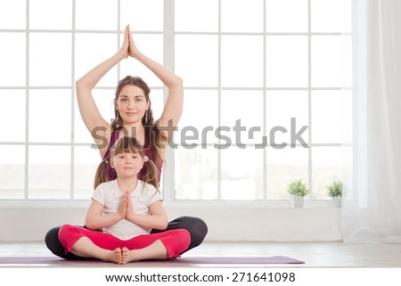 Young mother and daughter sitting in lotus position and doing yoga exercise in fitness studio with big windows on background