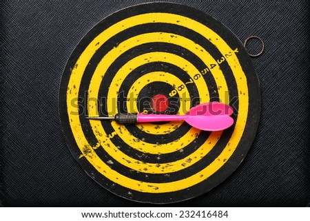 Used and vintage dartboard with arrow in the scene appear a lot of damaged hole from arrow marked put on the black color leather surface as a background represent the entertainment game.