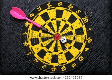 Used and vintage dartboard with arrow and dice in the scene appear a lot of damaged hole from arrow marked put on the black color leather surface as a background represent the entertainment game.