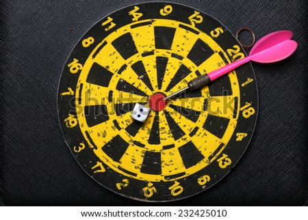 Used and vintage dartboard with arrow and dice in the scene appear a lot of damaged hole from arrow marked put on the black color leather surface as a background represent the entertainment game.