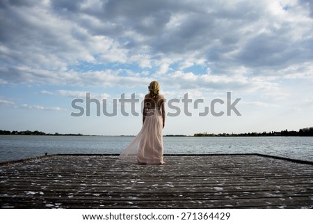 Back portrait of beautiful blond curly woman wearing evening peach color gown at lake.Fashionable and glamorous dress with chains.