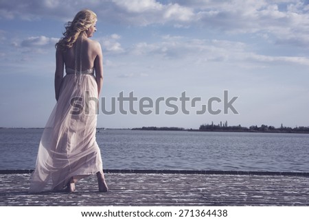 Barefoot blond woman wearing evening peach color gown standing on a deck with lake behind her while wind blows her gown.