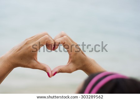 Woman making heart shape out of her hands looking towards the sea