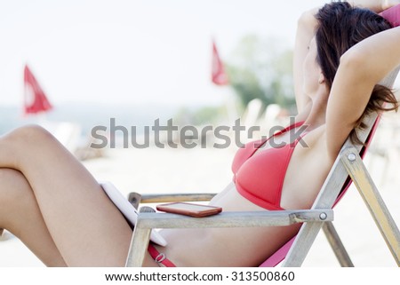 Good looking brunette woman wearing red bikini and using her tablet on a beach while sitting on a deck chair. She is looking away from the camera.