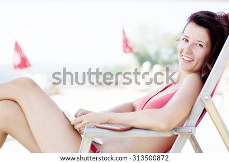 Good looking brunette woman wearing red bikini and using her tablet on a beach while sitting on a deck chair. She is smiling directly to camera.