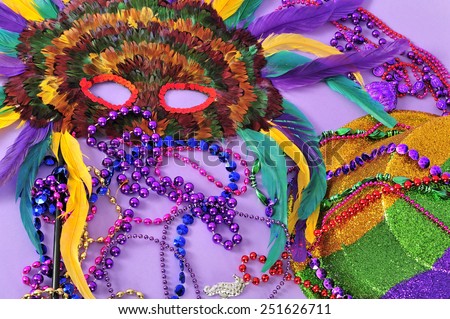 Mardi Gras feathered mask and top hat with Mardi Gras beads still life in gold green purple yellow and red on lilac background part of a series.