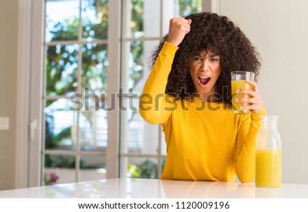 African american woman drinking healthy fruit juice at home annoyed and frustrated shouting with anger, crazy and yelling with raised hand, anger concept