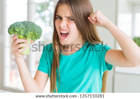 Beautiful young girl eating fresh broccoli annoyed and frustrated shouting with anger, crazy and yelling with raised hand, anger concept