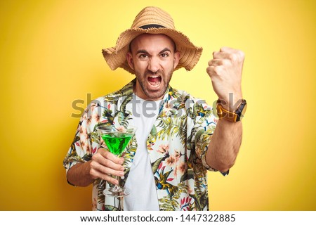 Young man wearing summer hawaiian flowers shirt and drinking a cocktail over yellow background annoyed and frustrated shouting with anger, crazy and yelling with raised hand, anger concept