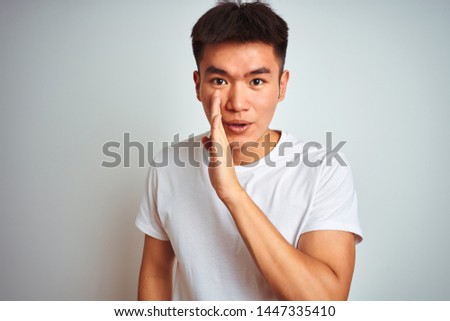 Young asian chinese man wearing t-shirt standing over isolated white background hand on mouth telling secret rumor, whispering malicious talk conversation
