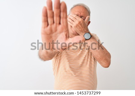 Homme de haut niveau aux cheveux gris portant un t-shirt rayé, debout sur fond blanc isolé, couvrant les yeux avec les mains et faisant un geste d